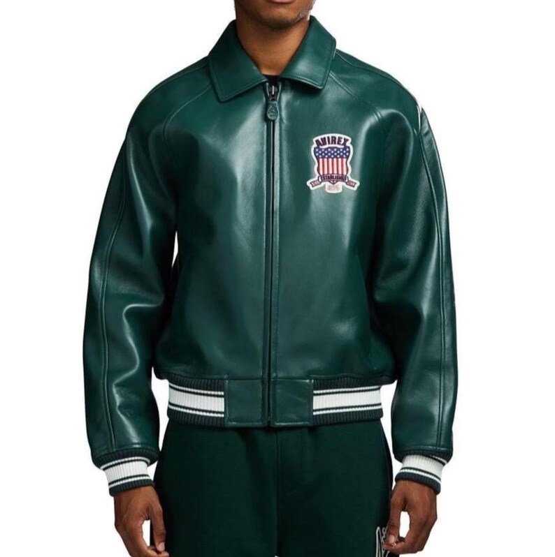 May include: A dark green leather jacket with a zip-up front and a collar. The jacket features a white and black striped band at the waist and cuffs. An embroidered patch with the text "Avirex" and a patriotic design is on the chest.