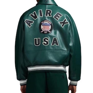 May include: A dark green leather jacket with the word "AVIREX" arched across the shoulders in white block letters. Below, the word "USA" is also in white. A circular patch with the words "ALL AMERICANS" and "ESTABLISHED 1975" is centered.