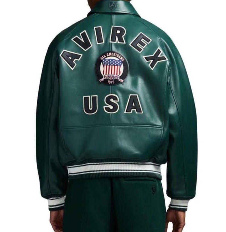 May include: A dark green leather jacket with the word "AVIREX" arched across the shoulders in white block letters. Below, the word "USA" is also in white. A circular patch with the words "ALL AMERICANS" and "ESTABLISHED 1975" is centered.
