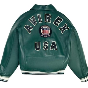 May include: A dark green leather jacket with white lettering that reads "AVIREX" and "USA". The jacket has a patch with the words "ALL AMERICANS ESTABLISHED 1975" and a design of the American flag. The jacket has a white and black striped trim.