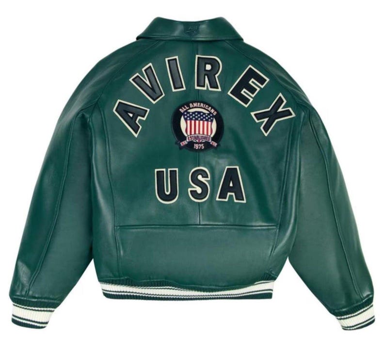 May include: A dark green leather jacket with white lettering that reads "AVIREX" and "USA". The jacket has a patch with the words "ALL AMERICANS ESTABLISHED 1975" and a design of the American flag. The jacket has a white and black striped trim.