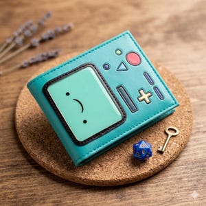Puede incluir: Cartera plegable de color turquesa con un diseño de cara de dibujos animados. La cartera tiene un borde negro alrededor de la cara y detalles de botones. También se ven una llave dorada, un dado azul y un posavasos de corcho.