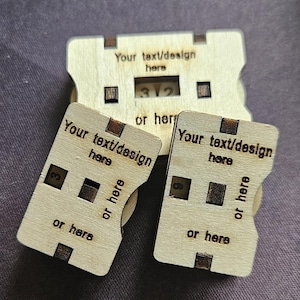 May include: Three wooden, laser-cut pieces with the text "Your text/design here" and "or here". One piece displays the number "312". The pieces are light brown with dark brown accents and are arranged on a purple surface.