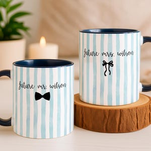Puede incluir: Dos tazas de cerámica con diseños de rayas azules y blancas. Una taza tiene un lazo negro y el texto "future mr. wilson". La otra taza tiene un lazo negro y el texto "future mrs. wilson". Ambas tazas tienen un interior y un asa de color azul oscuro.