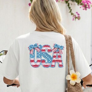 Puede incluir: Camiseta color marfil con la palabra "USA" en rojo, blanco y azul, con diseño de estrellas y rayas. La persona lleva la camiseta en un scooter. Una bolsa tejida con una flor cuelga a un lado.