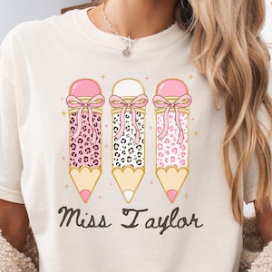Puede incluir: Una camiseta de manga corta de color crema con un gráfico de tres lápices con estampados de leopardo rosa y negro, cada uno adornado con un lazo rosa. Debajo de los lápices, el texto Miss Taylor está impreso en cursiva marrón oscuro.