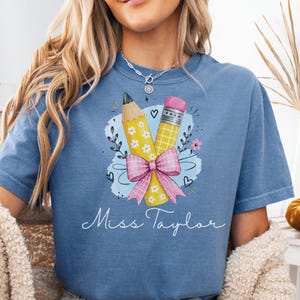 Puede incluir: Una camiseta azul de manga corta con un gráfico de dos lápices atados con un lazo de cuadros rosa, rodeados de pequeños detalles florales y una mancha de acuarela azul. El texto Miss Taylor está impreso en letra cursiva de color blanco.