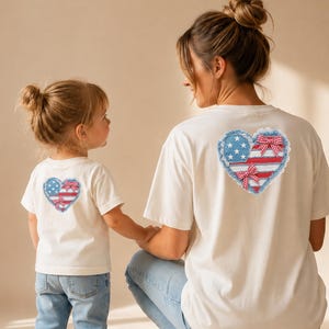Puede incluir: Dos camisetas blancas con diseños de corazón a juego con la bandera estadounidense. Las camisetas las llevan una madre y su hijo, con el niño agarrando la mano de la madre. El diseño incluye rayas rojas y blancas, estrellas azules y un ribete de encaje decorativo.