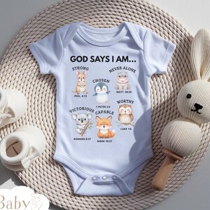 Puede incluir: Un body de bebé azul claro con el texto "GOD SAYS I AM..." e ilustraciones de animales. El body tiene las palabras "STRONG", "CHOSEN", "VICTORIOUS", "CAPABLE", "NEVER ALONE" y "WORTHY". Un sonajero de conejito blanco y botines de bebé también están en la imagen.