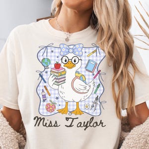 Puede incluir: Una camiseta color crema con un gráfico de un pato blanco con gafas y un lazo floral azul. El pato sostiene una pila de libros con una manzana encima. Rodeando al pato hay material escolar y el texto Miss Taylor escrito en color marrón.