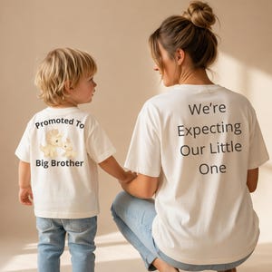Puede incluir: Dos camisetas blanquecinas con texto negro. Una dice "Promoted To Big Brother" con gráficos de dinosaurios. La otra dice "We're Expecting Our Little One." Ambas camisetas son usadas por un niño y un adulto.