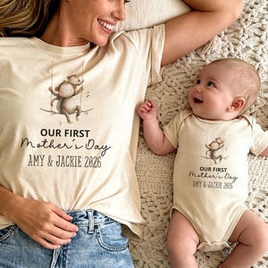 Puede incluir: Conjunto de camisetas y body beige a juego con un dibujo de un mono en un columpio. El texto "OUR FIRST Mother's Day AMY & JACKIE 2026" está impreso debajo. El adulto lleva vaqueros.