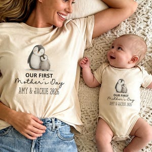 May include: Matching beige t-shirts and onesie featuring a penguin graphic. The text "OUR FIRST Mother's Day" is printed below the penguin design, along with the names "AMY & JACKIE 2026". The adult is wearing a t-shirt and the baby is wearing a onesie.