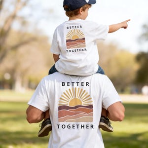May include: Two white short-sleeve t-shirts featuring a graphic on the back with a sun rising over layered hills in shades of tan, brown, and orange. The text "BETTER TOGETHER" is printed in black capital letters above and below the graphic.