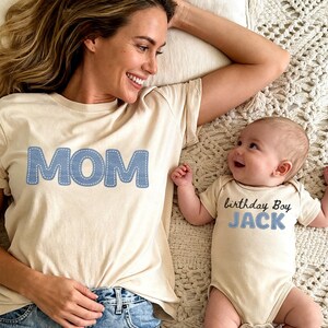 Puede incluir: Una camiseta color crema con la palabra MOM en letras azules con un contorno cosido en blanco, y un body de bebé a juego color crema con el texto birthday Boy JACK en letras negras y azules con un contorno cosido en blanco.