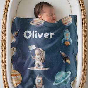 Puede incluir: Una manta para bebé de color azul oscuro con un tema espacial que incluye planetas, cohetes y un astronauta sosteniendo una bandera estadounidense. El nombre Oliver está impreso en letras blancas en la parte superior con cuerpos celestes.