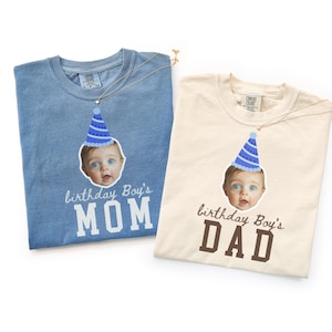 Puede incluir: Una camiseta azul con el texto birthday Boy's MOM y una camiseta crema con el texto birthday Boy's DAD. Ambas camisetas presentan un gráfico de un bebé con un gorro de fiesta azul. Las camisetas tienen una etiqueta Comfort Colors en el cuello.