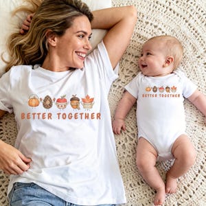 Puede incluir: Una camiseta blanca de manga corta y un body blanco de manga corta para bebé, ambos con un gráfico de una calabaza, piña, manzana, bellota y hoja de arce en tazas con el texto BETTER TOGETHER impreso en color naranja debajo.