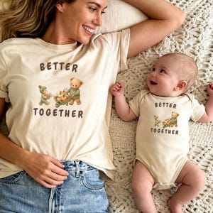 Puede incluir: Una camiseta de color crema y un body de bebé a juego, ambos con un gráfico de dos ositos de peluche con una carretilla de calabazas y el texto "BETTER TOGETHER" en letras marrones. Las prendas están hechas de una tela de color claro.