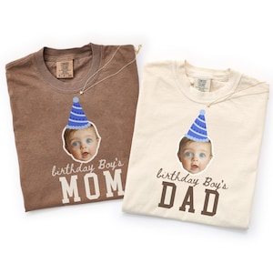 Puede incluir: Dos camisetas de manga corta, una marrón y otra crema, cada una con el gráfico de un bebé con un gorro de fiesta azul. La camiseta marrón tiene el texto "birthday Boy's MOM" y la crema tiene el texto "birthday Boy's DAD" impreso debajo.