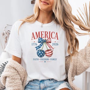 Puede incluir: Camiseta blanca con la palabra "AMERICA" en rojo y los años 1776 y 2026. La camiseta presenta un diseño de cerezas con un lazo y las palabras "FAITH FREEDOM FAMILY".