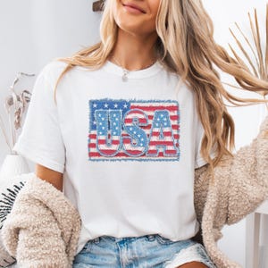Puede incluir: Camiseta blanca con un diseño patriótico "USA". Las letras están rellenas con las estrellas y rayas de la bandera estadounidense, dentro de un marco de estilo denim. El diseño celebra el orgullo nacional estadounidense.
