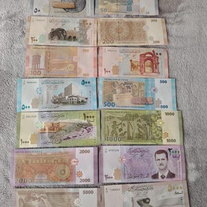 May include: A collection of Syrian banknotes, featuring denominations from 50 to 5000 Syrian pounds. The bills display various designs, including historical landmarks, portraits, and intricate patterns. The banknotes are arranged on a neutral-colored surface.