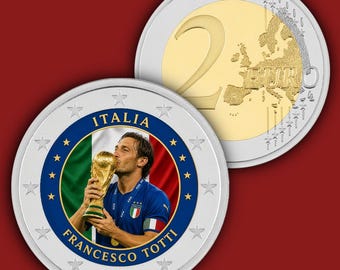 May include: A commemorative two-euro coin featuring a portrait of Francesco Totti kissing the World Cup trophy against an Italian flag background. The text reads ITALIA and FRANCESCO TOTTI, surrounded by stars on a silver and gold metallic surface.