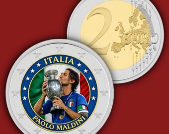 May include: A two-euro coin featuring a portrait of Paolo Maldini kissing a trophy against the Italian flag. The coin has a silver outer ring with stars and a central image surrounded by the text ITALIA and PAOLO MALDINI in blue.