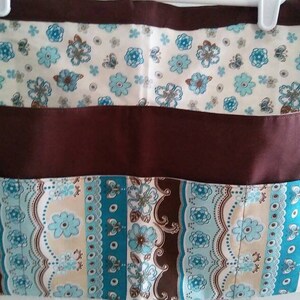 May include: A brown and blue patterned apron with three pockets. The apron has a white top section with a floral pattern, a brown middle section, and a blue and brown bottom section with a paisley pattern.