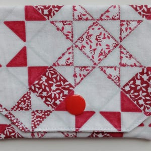 Puede incluir: Un monedero de tela blanca y roja con un diseño de patchwork. El patrón presenta triángulos rojos y cuadrados florales, con un cierre de botón rojo. El monedero tiene forma rectangular.