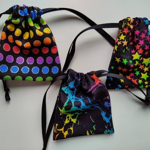 May include: Three drawstring bags with black fabric and colorful patterns. The first bag has a rainbow polka dot pattern, the second bag has a rainbow splatter pattern, and the third bag has a rainbow star pattern.