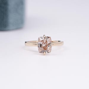 Peut inclure: Une bague en or ornée d'une pierre précieuse couleur champagne taille coussin, fixée par quatre griffes. L'anneau est lisse et poli, présentant une teinte métallique chaude qui complète la pierre à facettes au centre du bijou.