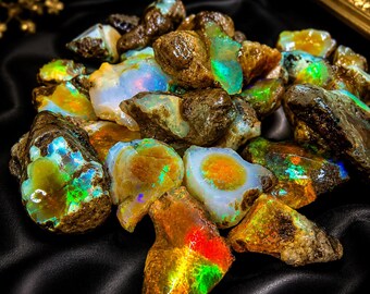 May include: A collection of rough opal gemstones, showcasing a variety of colors including green, blue, red, and yellow. The opals have a rough, natural texture and are displayed on a black surface. The gemstones are a mix of sizes and shapes.