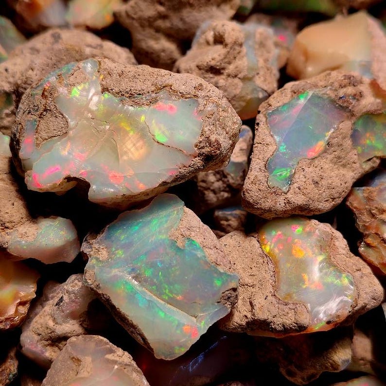 May include: A collection of rough opal stones, showcasing a play of color. The opals display iridescent flashes of green, blue, red, and yellow within a matrix of brown rock. The stones vary in size and shape, creating a natural, earthy aesthetic.