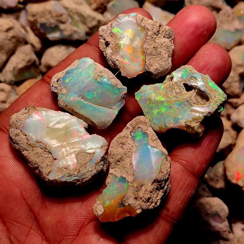 May include: Five raw opal gemstones with vibrant iridescent colors of green, blue, orange, and yellow. The opals are set in a rough, brown matrix. The gemstones are held in a hand.