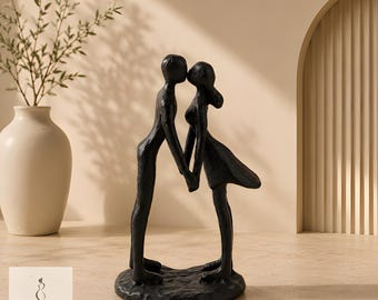 May include: A black sculpture of two figures standing face to face, holding hands. The figures have smooth, elongated forms and are mounted on a textured, circular base. The piece features a matte finish and a stylized, abstract design.
