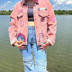 Puede incluir: Una chaqueta de mezclilla rosa polvoriento adornada con varios parches, incluyendo un logo de Nike Air, corazones y diseños de arcoíris, con botones de metal plateado y cuello. La chaqueta se lleva sobre un top negro y jeans azul claro con aberturas laterales.