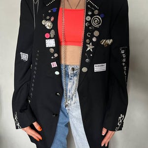 May include: Black blazer adorned with various pins, patches, and chains. The blazer features decorative stitching and the text "BLACKROLL monobrand". The model is wearing a neon orange top and blue jeans. A silver chain necklace hangs down the front.