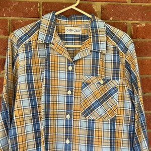 May include: A long-sleeved plaid shirt in blue, yellow, and white with a button-up front and a single chest pocket. The shirt has the text "CABIN CREEK" on the label.