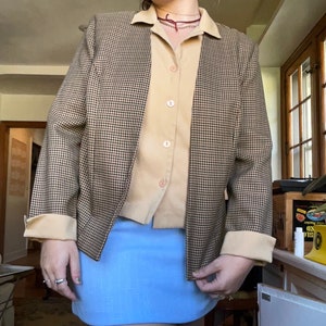 May include: A tan button-down shirt and a light blue skirt are worn under a brown and black houndstooth blazer. The blazer has tan cuffs and a tan collar. The outfit is a mix of textures and colors.