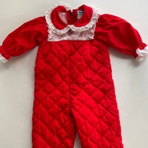Könnte beinhalten: Roter, gesteppter Baby-Overall mit langen Ärmeln und weißem Spitzenkragen. Der Overall hat ein Rautenmuster und Knopfverschlüsse am unteren Ende. Die Ärmel und der Kragen sind mit weißer Spitze verziert.