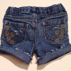 May include: Blue denim shorts with distressed details and rolled cuffs. The back pockets are adorned with embroidered floral designs. The shorts have a classic five-pocket design and a button and zipper closure.