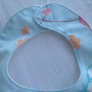 May include: A light blue baby bib with a white trim and a colorful pattern of stars, hearts, and balloons. The bib has a hook and loop closure.