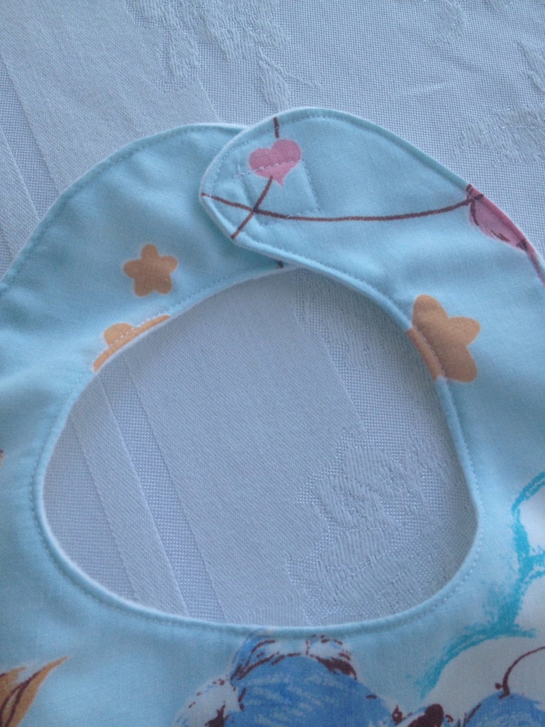 May include: A light blue baby bib with a white trim and a colorful pattern of stars, hearts, and balloons. The bib has a hook and loop closure.