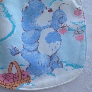 May include: A blue Care Bear with a cloud on its belly and a basket of hearts. The Care Bear is holding a heart in its paw and is surrounded by clouds and hearts. The background is light blue.