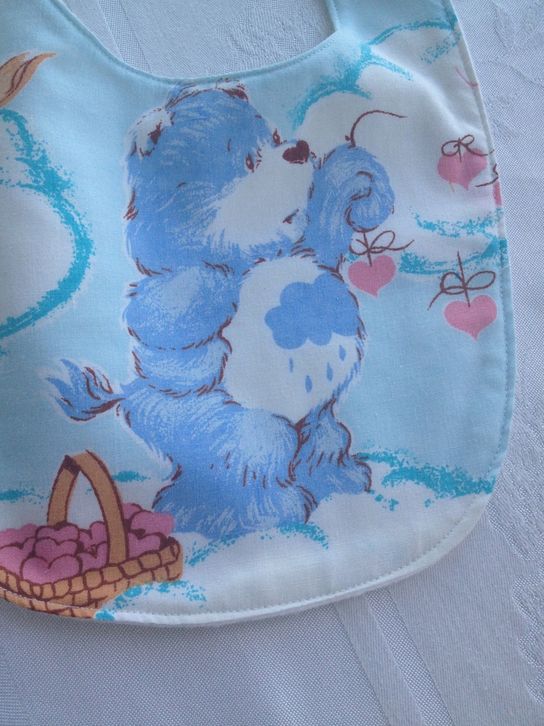May include: A blue Care Bear with a cloud on its belly and a basket of hearts. The Care Bear is holding a heart in its paw and is surrounded by clouds and hearts. The background is light blue.