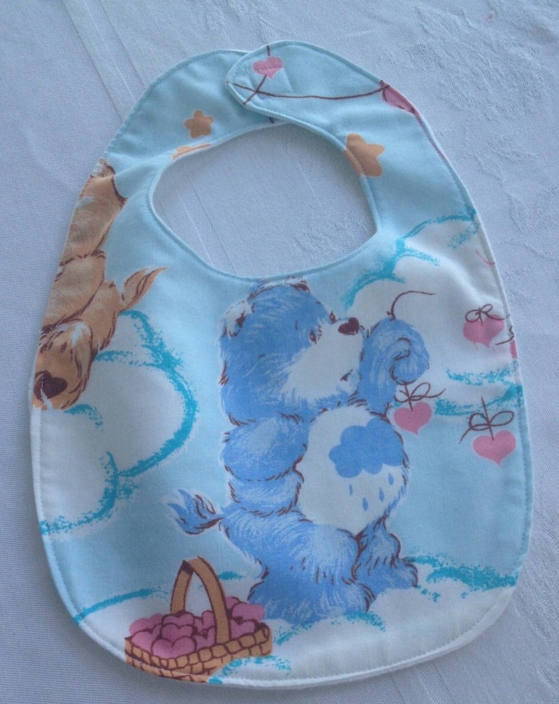 May include: A blue baby bib with a Care Bear design. The Care Bear is holding a basket of hearts and there are hearts and stars in the background.