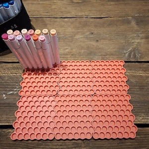 May include: A selection of art markers in a black case, alongside coral-coloured, interlocking silicone mats. The markers are various shades of pink, peach, and beige. The mats feature a honeycomb design and are arranged on a dark wooden surface.