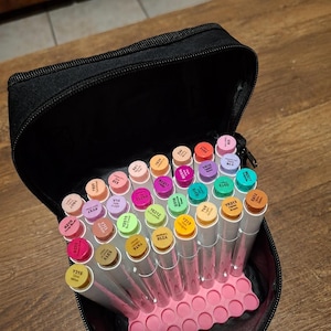 Puede incluir: Un estuche negro con cremallera lleno de una colección de marcadores multicolores. Los marcadores están dispuestos verticalmente en una base de rejilla de plástico rosa, cada uno con una tapa de color con un código alfanumérico impreso.